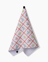 The Fall Flowers tea towel by Claire Iglesias features a red checkered diamond pattern with blue, yellow, and red floral designs. Made from recycled materials, it hangs by a black loop against a plain white background.