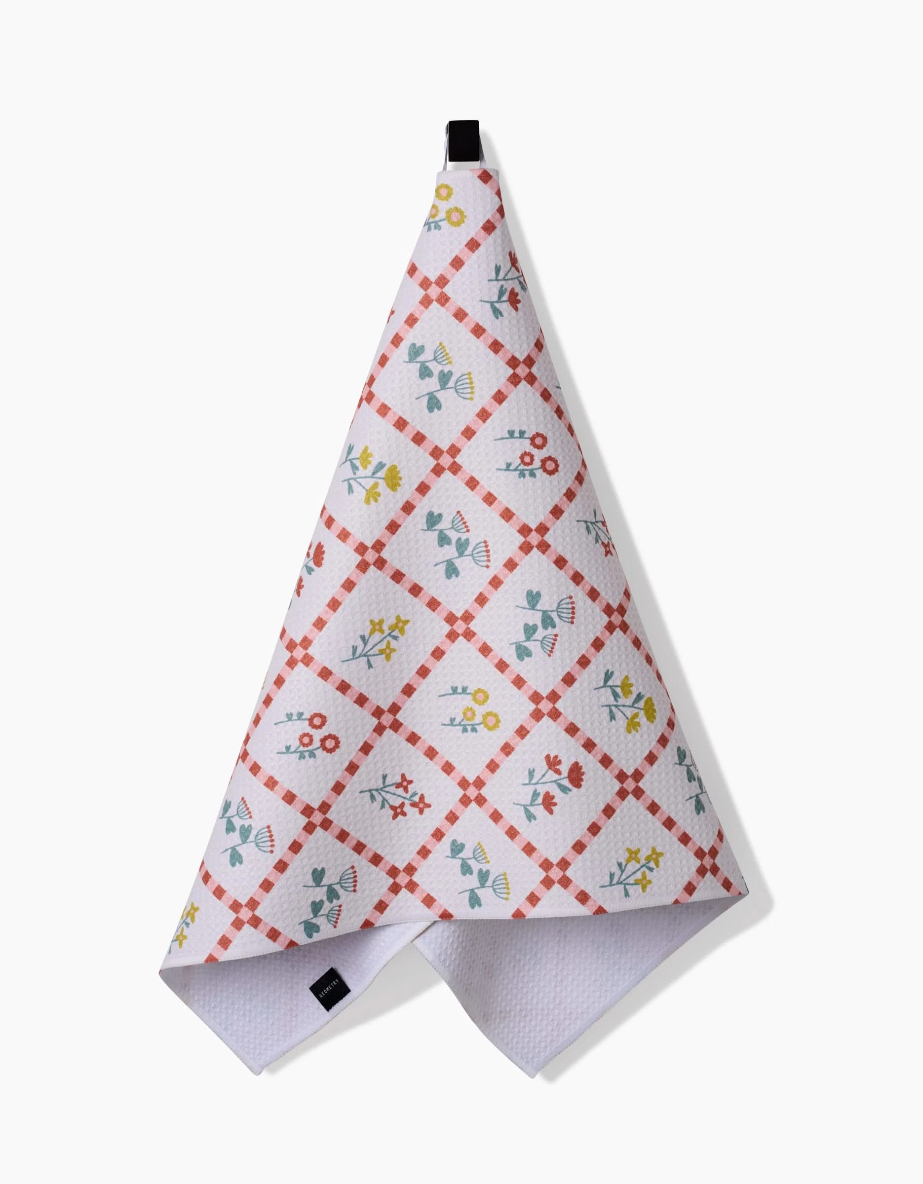 The Fall Flowers tea towel by Claire Iglesias features a red checkered diamond pattern with blue, yellow, and red floral designs. Made from recycled materials, it hangs by a black loop against a plain white background.