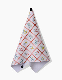 The Fall Flowers tea towel by Claire Iglesias features a red checkered diamond pattern with blue, yellow, and red floral designs. Made from recycled materials, it hangs by a black loop against a plain white background.
