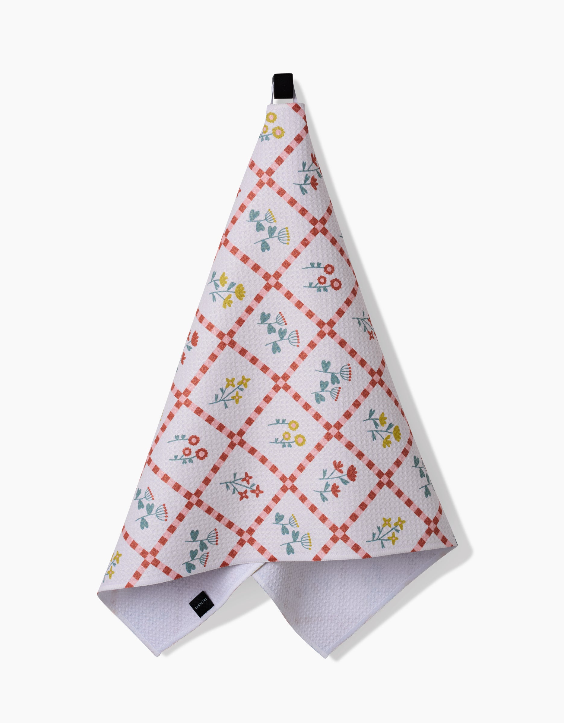 The Fall Flowers tea towel by Claire Iglesias features a red checkered diamond pattern with blue, yellow, and red floral designs. Made from recycled materials, it hangs by a black loop against a plain white background.