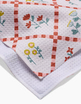 A close-up of Claire Iglesias Fall Flowers kitchen towel, ultra-absorbent with a red checkered pattern and colorful yellow, red, and blue floral designs. The folded tea towel shows both printed and plain white textured sections.