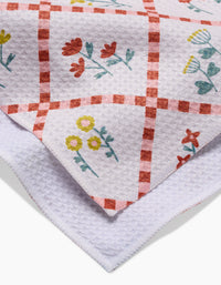 A close-up of Claire Iglesias Fall Flowers kitchen towel, ultra-absorbent with a red checkered pattern and colorful yellow, red, and blue floral designs. The folded tea towel shows both printed and plain white textured sections.