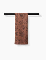 The Fall Foliage kitchen towel by Aubrey Fairchild, featuring a black floral and leaf pattern on a reddish-brown, eco-friendly fabric, hangs neatly from a simple black towel bar against a white background.
