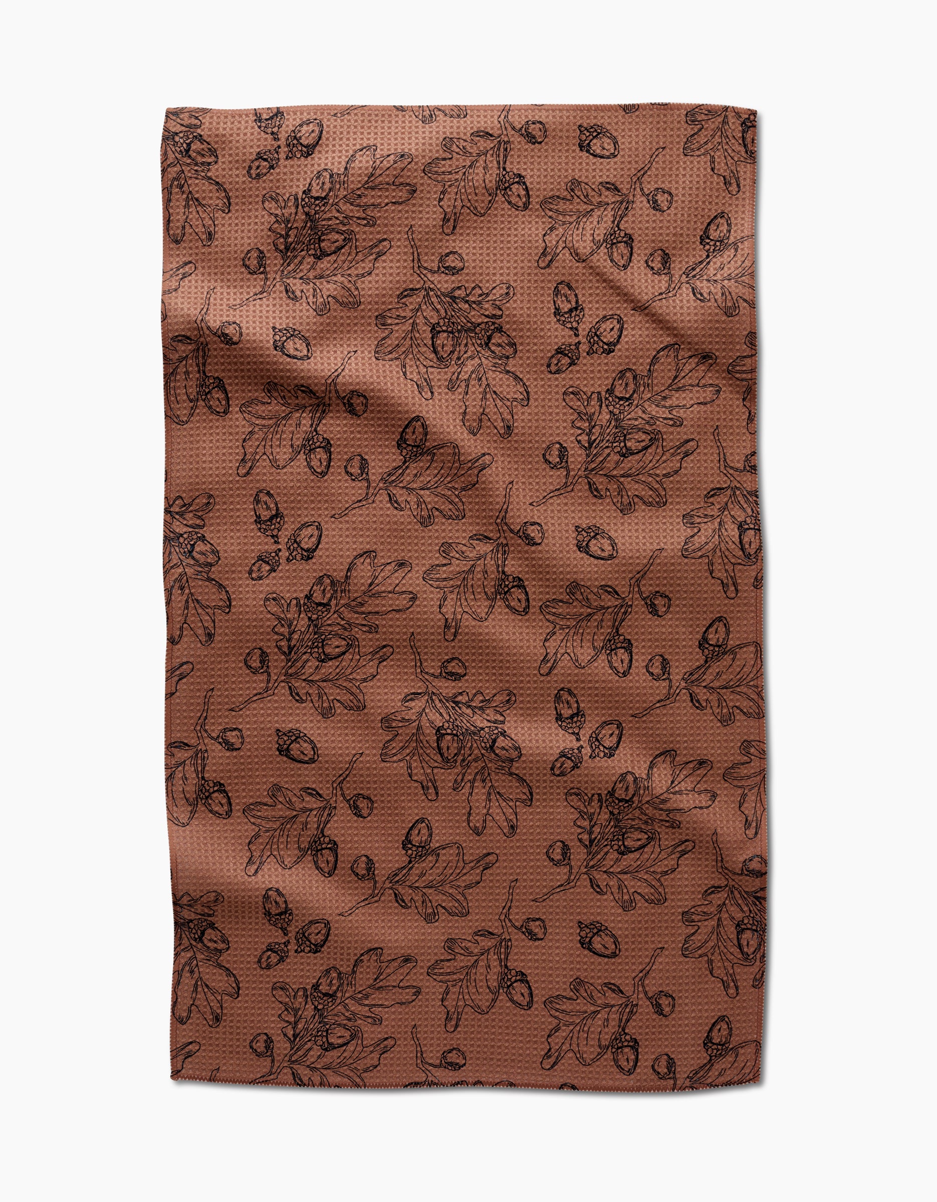 The Fall Foliage kitchen towel by Aubrey Fairchild features eco-friendly brown fabric with a black oak leaf and acorn pattern, creating an autumnal, botanical vibe. Laid flat with slight wrinkles, it makes a charming addition to any kitchen.