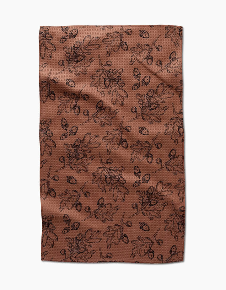 The Fall Foliage kitchen towel by Aubrey Fairchild features eco-friendly brown fabric with a black oak leaf and acorn pattern, creating an autumnal, botanical vibe. Laid flat with slight wrinkles, it makes a charming addition to any kitchen.