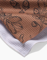 Aubrey Fairchilds Fall Foliage tea towel features textured brown fabric with black botanical sketches of leaves, acorns, and berries, shown overlapping a white textured cloth.