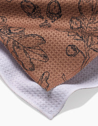 Aubrey Fairchilds Fall Foliage tea towel features textured brown fabric with black botanical sketches of leaves, acorns, and berries, shown overlapping a white textured cloth.