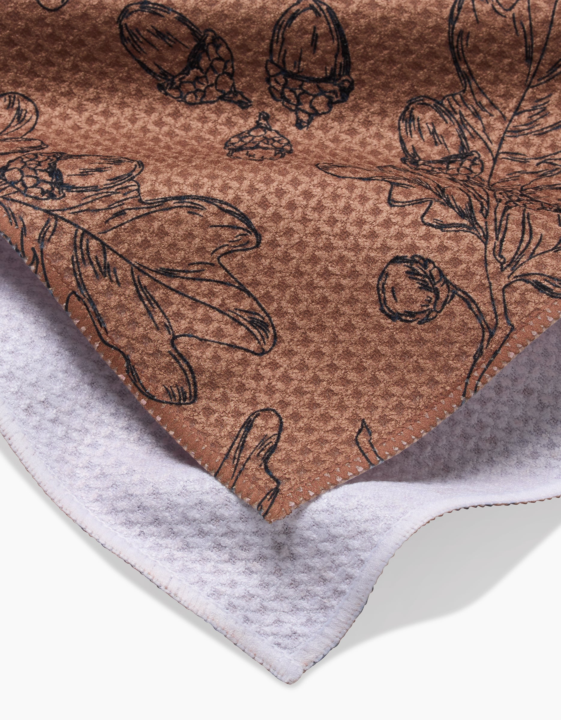 Aubrey Fairchilds Fall Foliage tea towel features textured brown fabric with black botanical sketches of leaves, acorns, and berries, shown overlapping a white textured cloth.