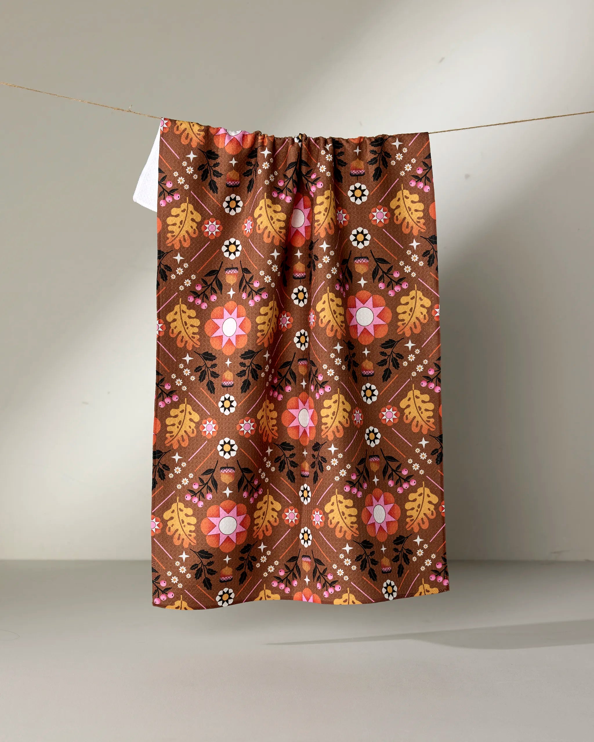 The "Fall Folk" tea towel by Megan Roy features a colorful floral and geometric pattern in pink, orange, yellow, and white on brown, hanging against a neutral background.