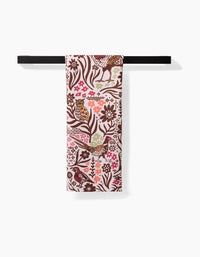 The Fall Fowl tea towel by Kayla Ann hangs on a black rod, showcasing a vibrant bird, flower, and leaf pattern in brown, pink, red, and green on a light background—made from recycled materials.