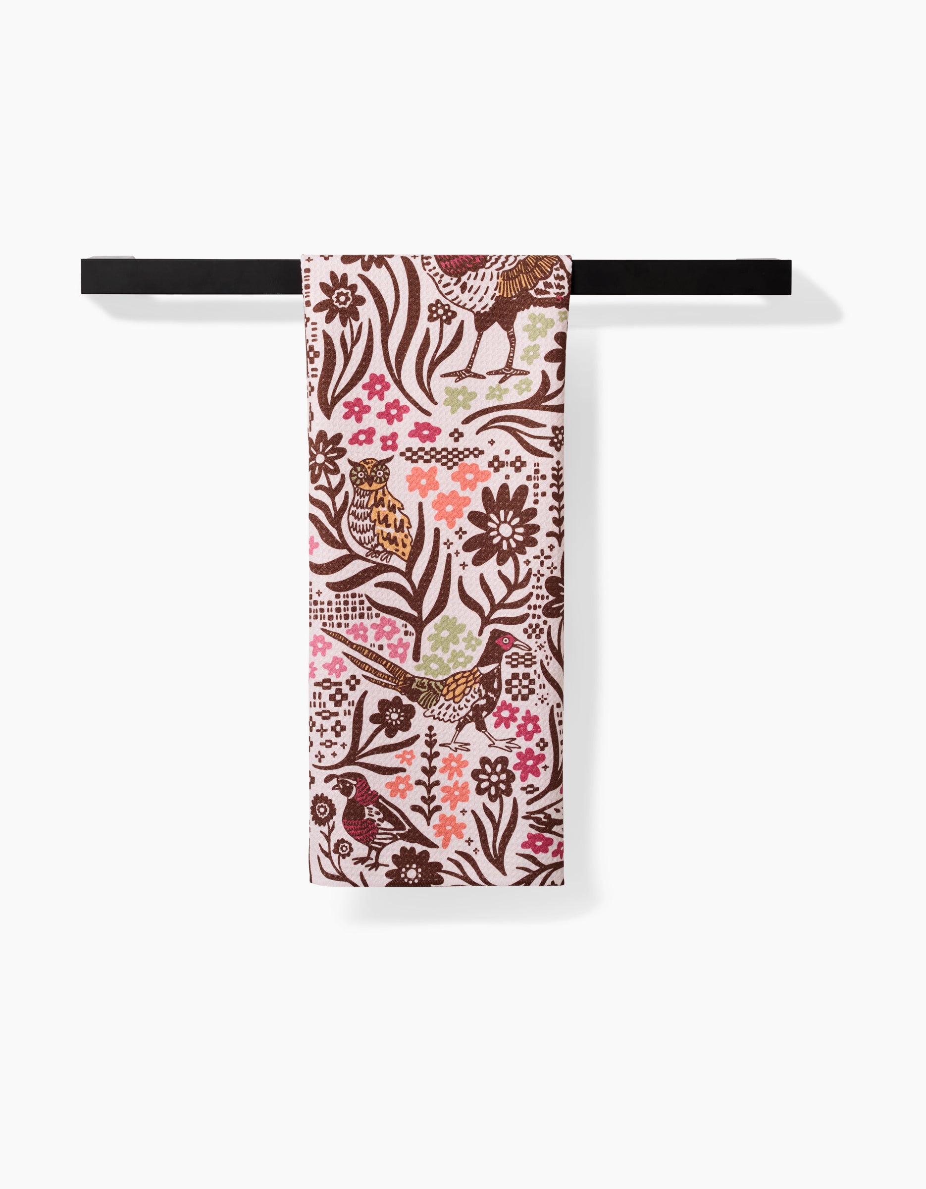 The Fall Fowl tea towel by Kayla Ann hangs on a black rod, showcasing a vibrant bird, flower, and leaf pattern in brown, pink, red, and green on a light background—made from recycled materials.