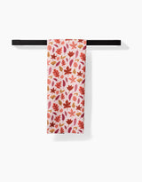 The Kayla Ann Fall Leaves kitchen towel, featuring an eco-friendly white fabric with red, pink, and gold autumn leaves, is neatly folded on a simple black towel bar against a plain white background.