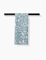 The Claire Iglesias Fall Mushrooms And Leaves green and white kitchen tea towel, featuring a leafy and mushroom pattern and made from recycled materials, hangs neatly on a black towel bar against a white background.