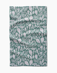 The Fall Mushrooms And Leaves tea towel by Claire Iglesias features a green background with white patterns of leaves, acorns, mushrooms, and botanical elements, shown with visible folds and creases.