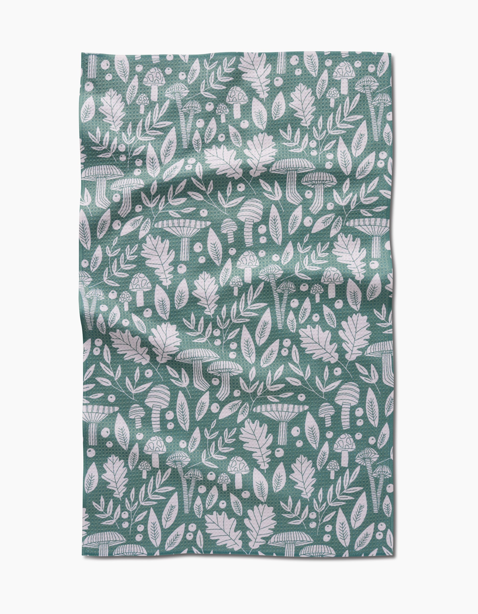 The Fall Mushrooms And Leaves tea towel by Claire Iglesias features a green background with white patterns of leaves, acorns, mushrooms, and botanical elements, shown with visible folds and creases.