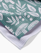 A close-up of the Claire Iglesias Fall Mushrooms And Leaves tea towel, featuring a teal recycled fabric with a white botanical and mushroom print, layered over a white waffle-textured cloth.