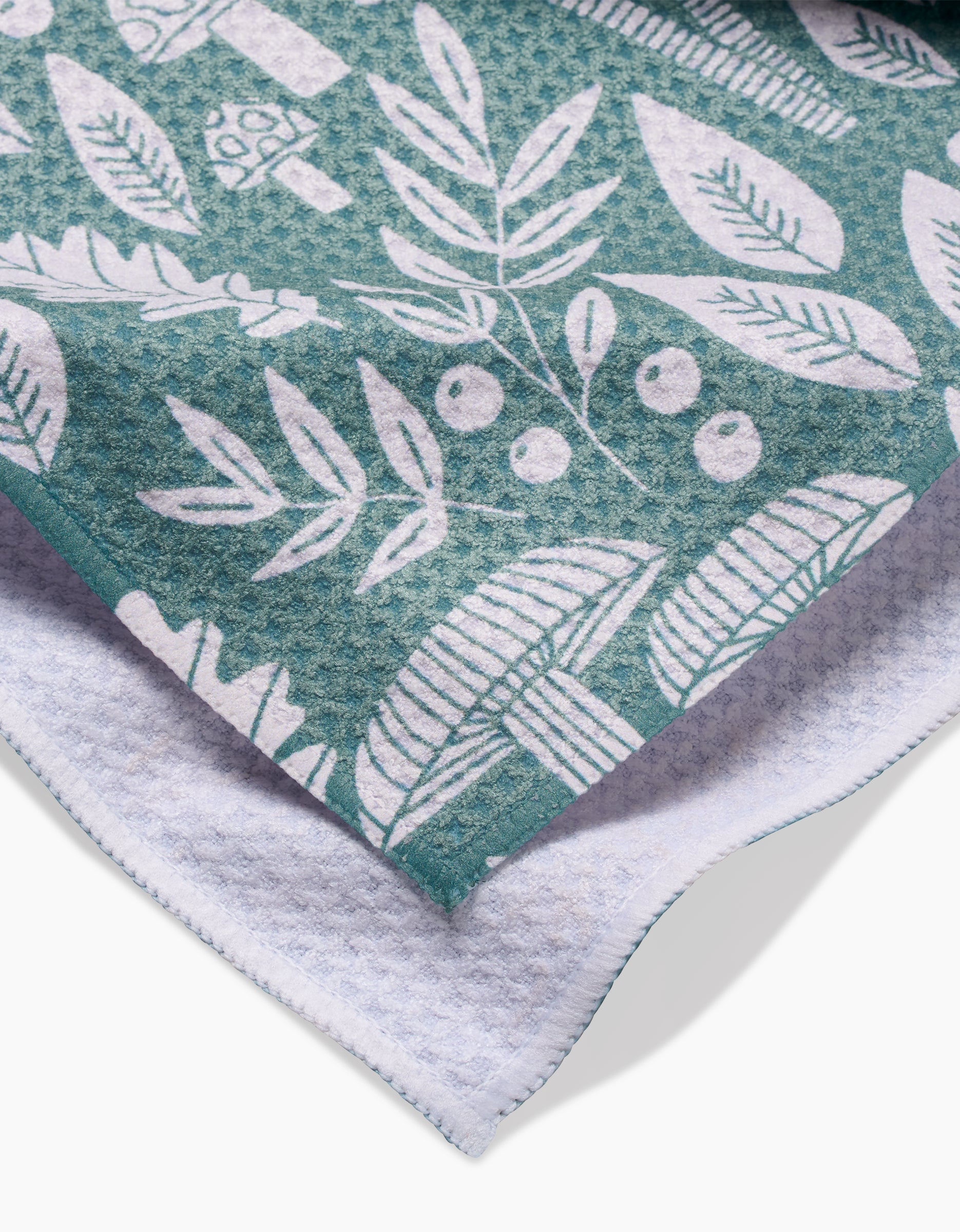 A close-up of the Claire Iglesias Fall Mushrooms And Leaves tea towel, featuring a teal recycled fabric with a white botanical and mushroom print, layered over a white waffle-textured cloth.