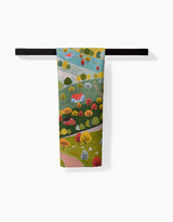 The Fall Sunflower Cottage towel by Ceyda Alasar features a whimsical landscape with hills, houses, cows, and trees. Ultra-absorbent and colorful, it’s shown hanging on a black towel bar against a white background.