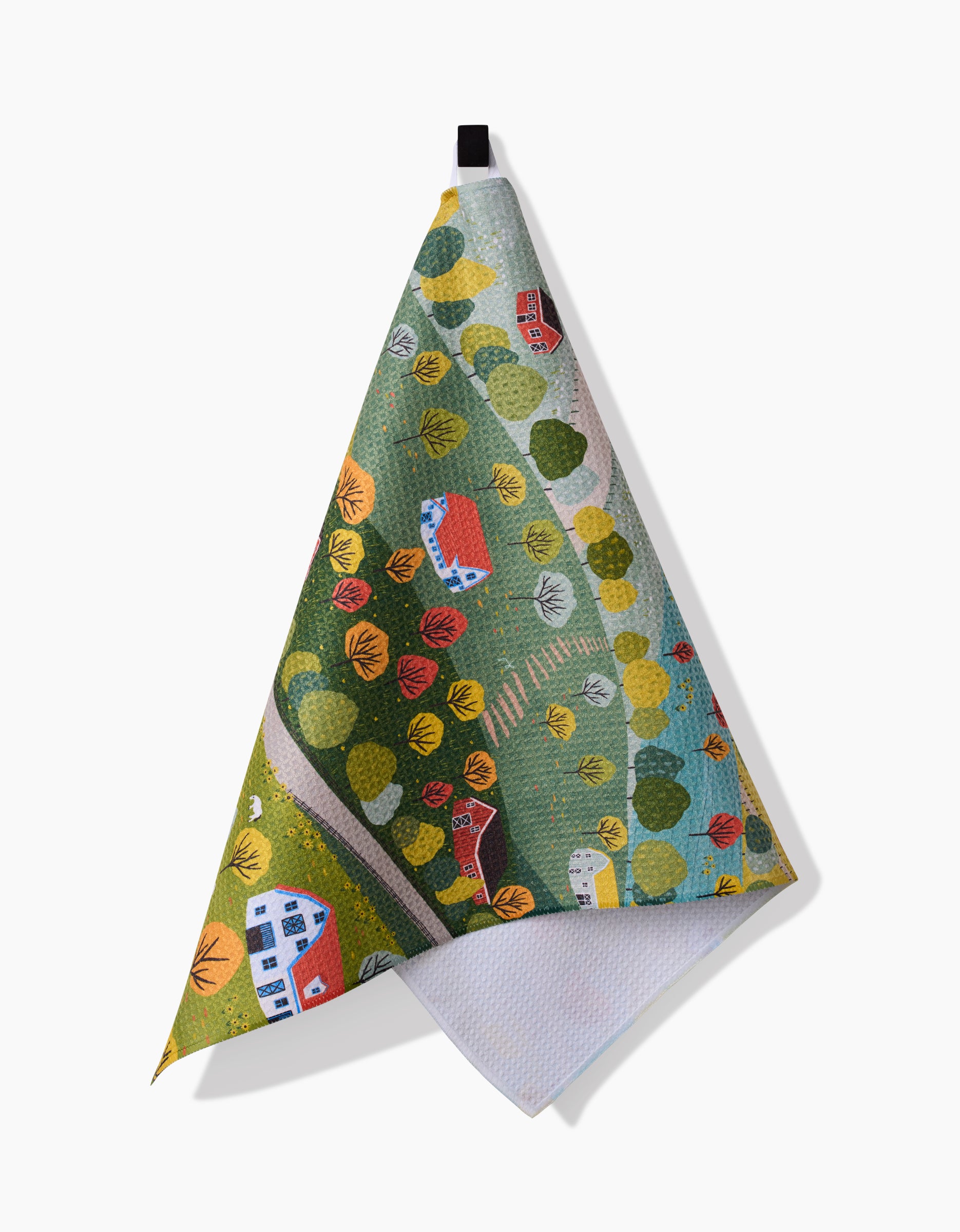 The Fall Sunflower Cottage tea towel by Ceyda Alasar is made from recycled materials and features a vibrant landscape with trees, paths, and cottages in red, blue, and white, hanging by a black loop against a white background.