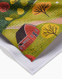 A close-up of the Fall Sunflower Cottage tea towel by Ceyda Alasar, featuring a colorful illustration of a red barn, autumn trees, and green fields, with the white textured underside partly visible.