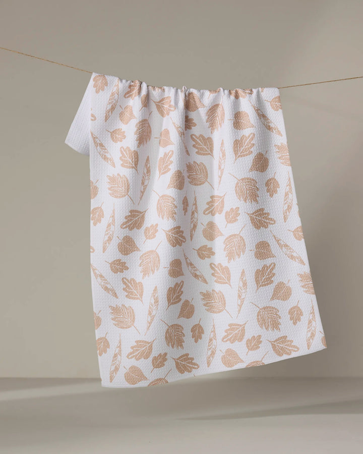 The Fallen Leaf tea towel by Hufton Studio, featuring an eco-friendly white fabric with a light brown leaf pattern, hangs on a clothesline against a neutral background.
