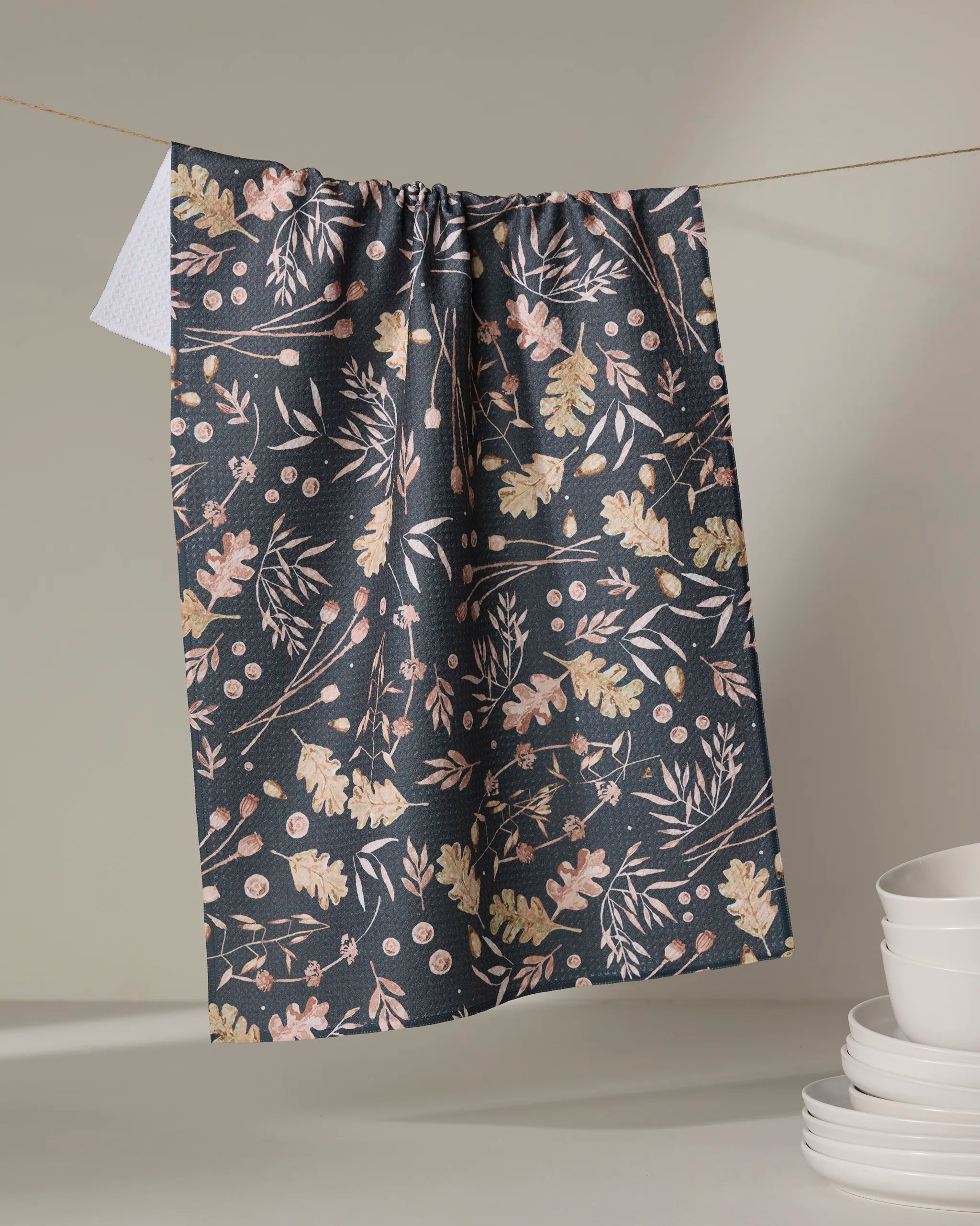 The Fallen Oak kitchen towel by Katie O'Shea, featuring a pastel botanical print on a dark background and made from recycled materials, hangs above neatly stacked white bowls and plates.