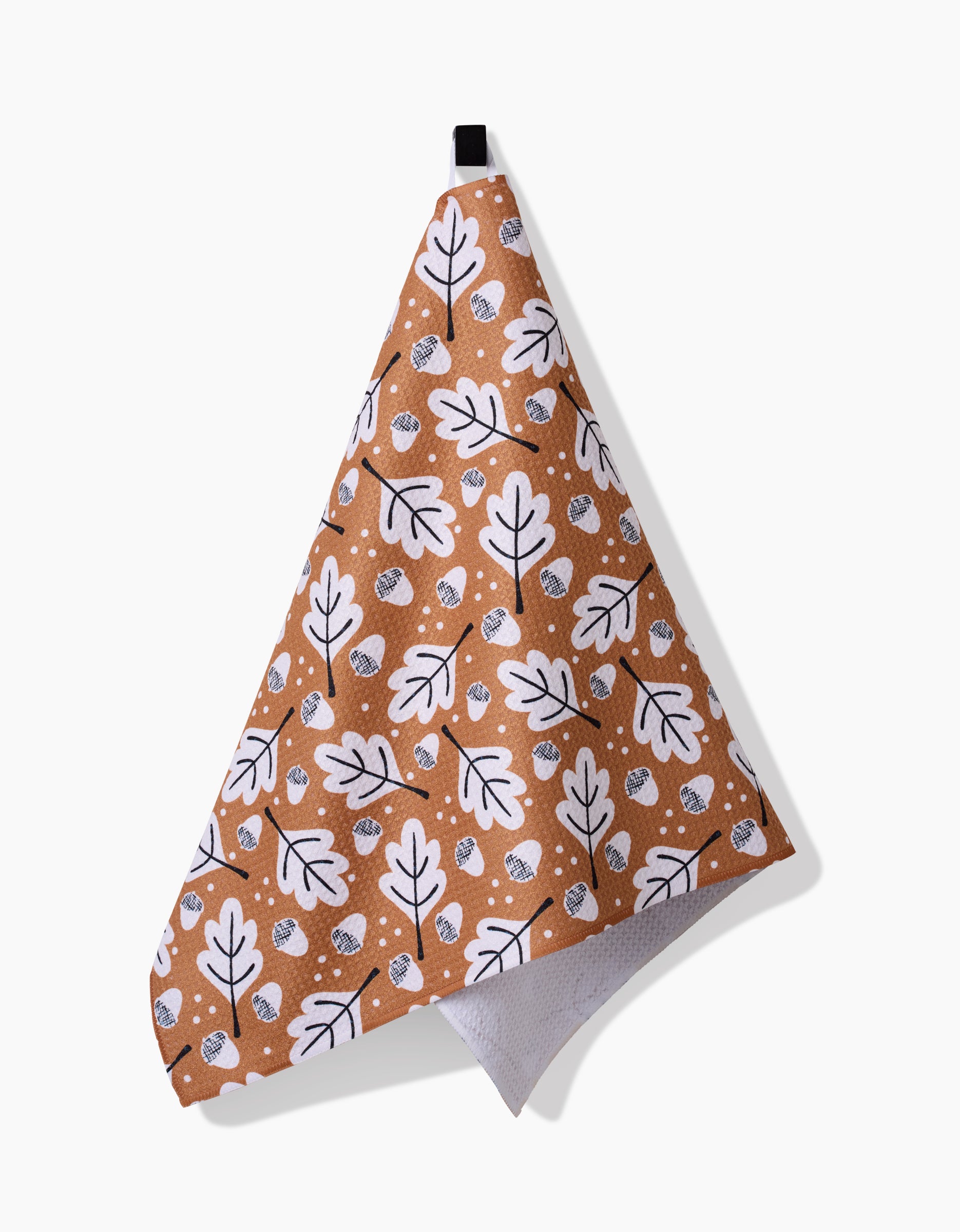 The Falling Acorns tea towel by Charlie Rowan Designs, made from Geoweave fabric, features a white and black leaf and acorn pattern on brown and hangs from a black loop against a white background.