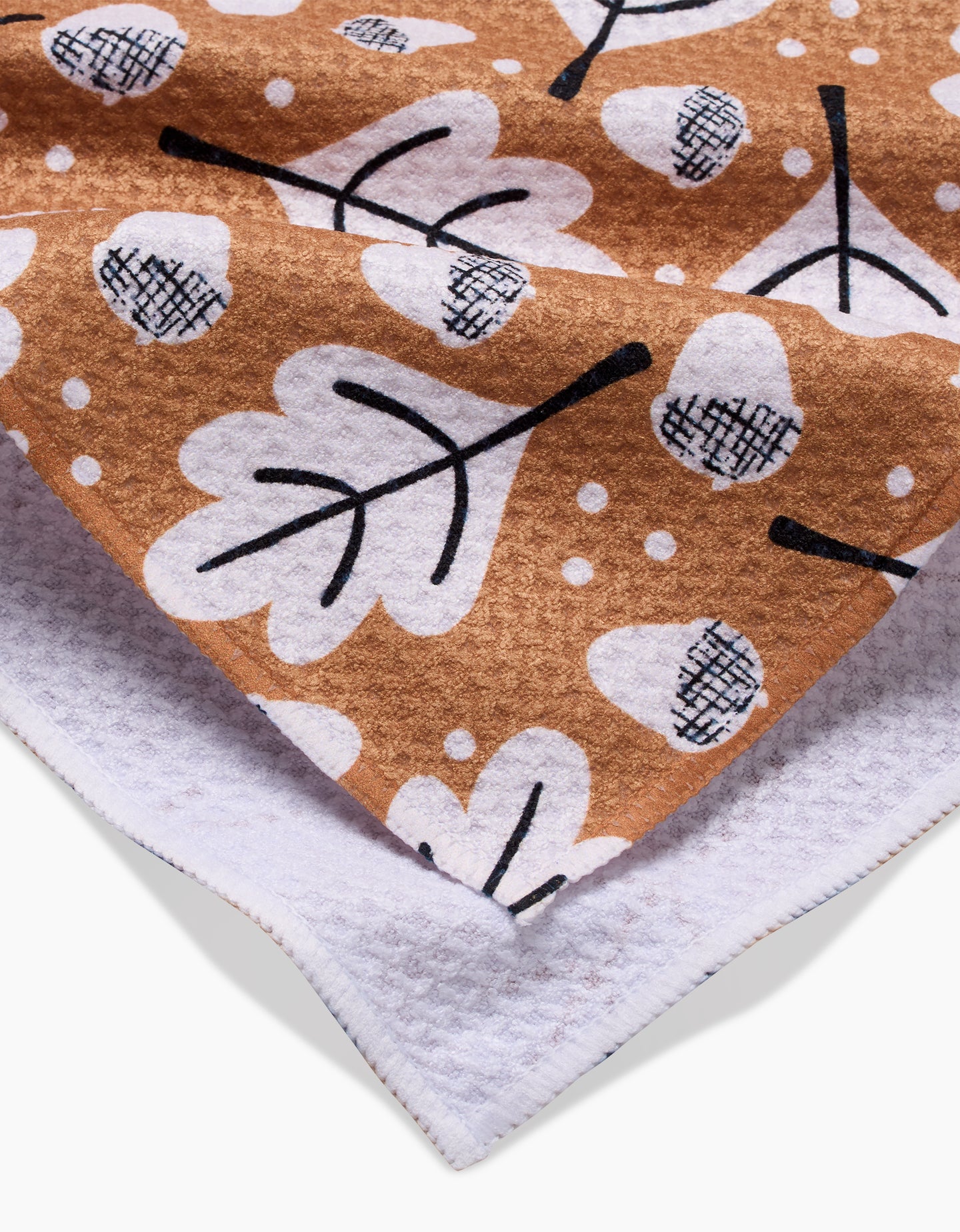 A close-up of a Charlie Rowan Designs Falling Acorns tea towel, made from Geoweave fabric, featuring a brown base with white leaves and dots, black stems, and a textured white underside.