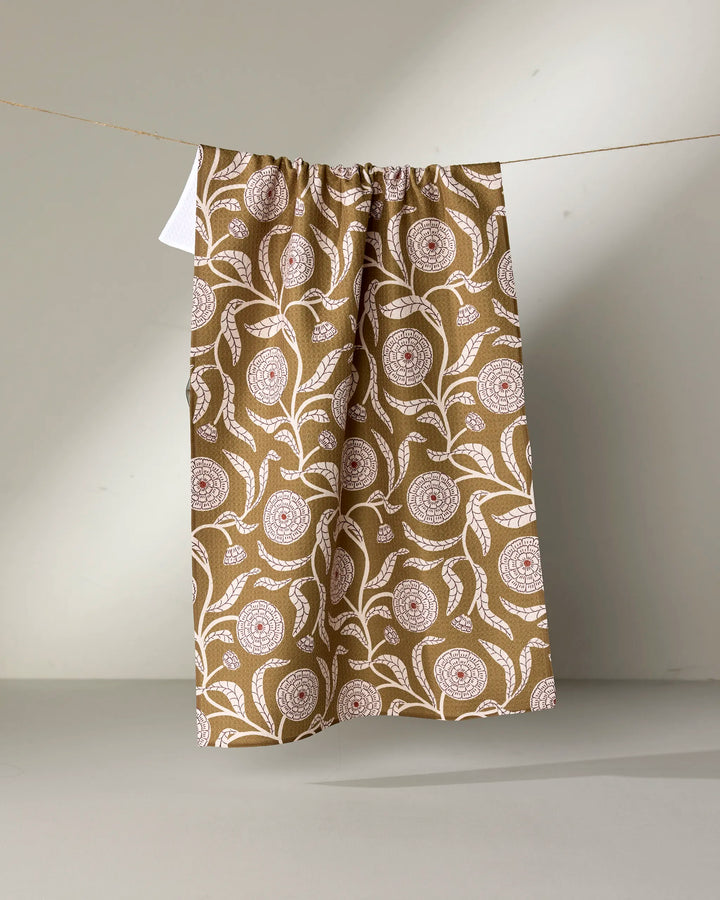 The Falling Flowers tea towel by Teri the Butter, featuring a white and red floral pattern on brown fabric, hangs on a string against a light gray background.