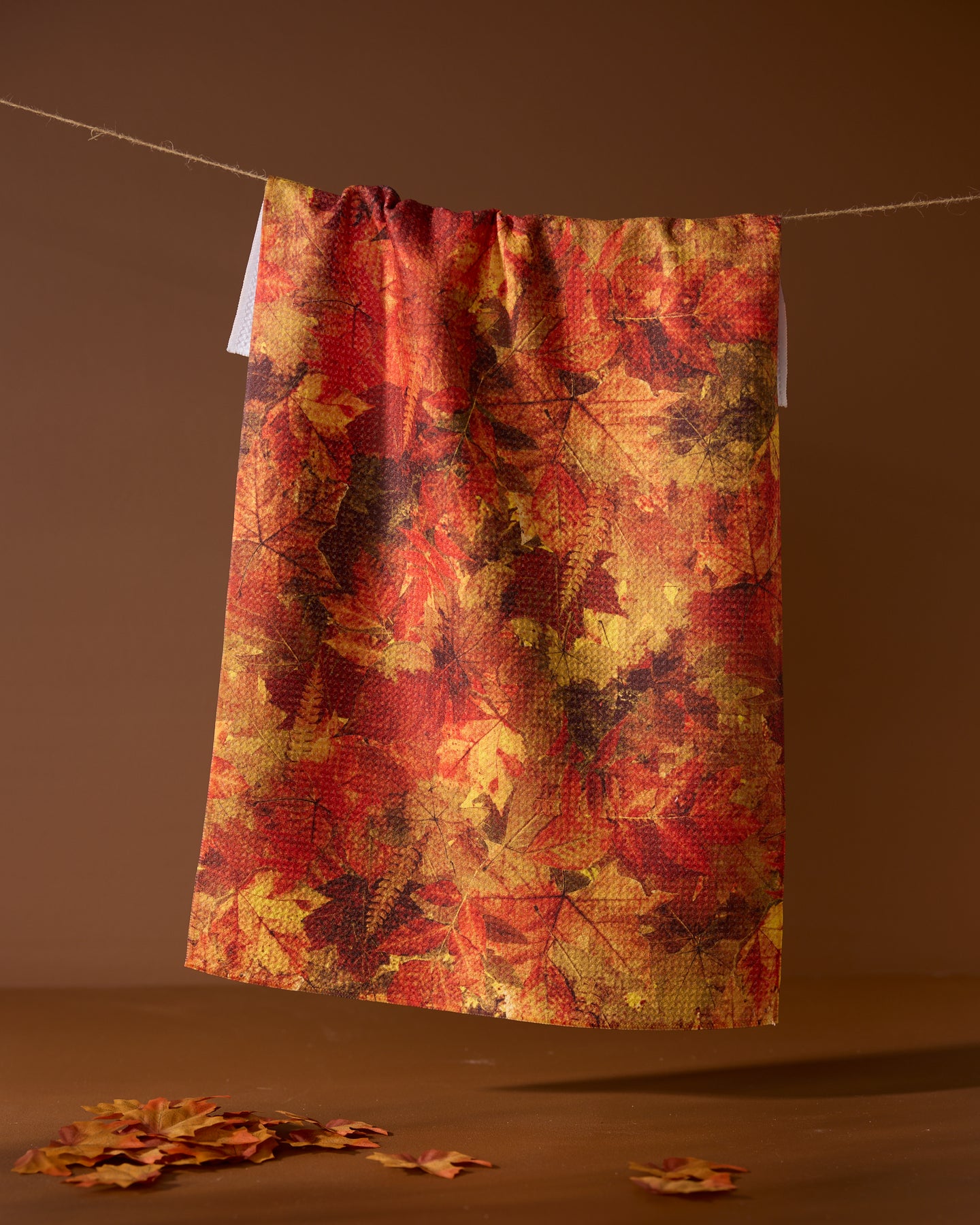 The Geometry Falling Foliage tea towel, featuring an autumn leaf pattern in red, orange, and yellow, hangs on a string against a brown background with scattered leaves below.