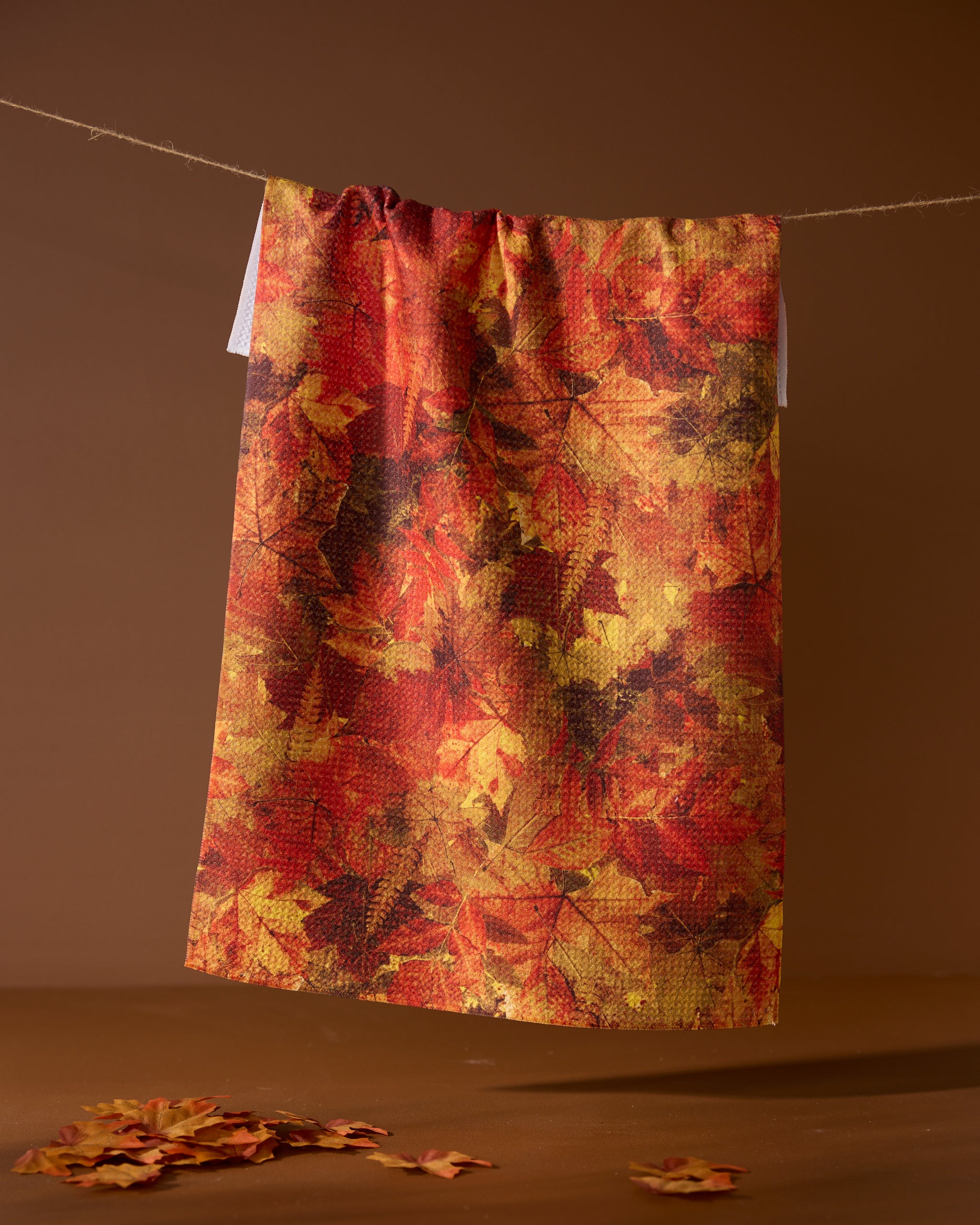 The Geometry Falling Foliage tea towel, featuring an autumn leaf pattern in red, orange, and yellow, hangs on a string against a brown background with scattered leaves below.