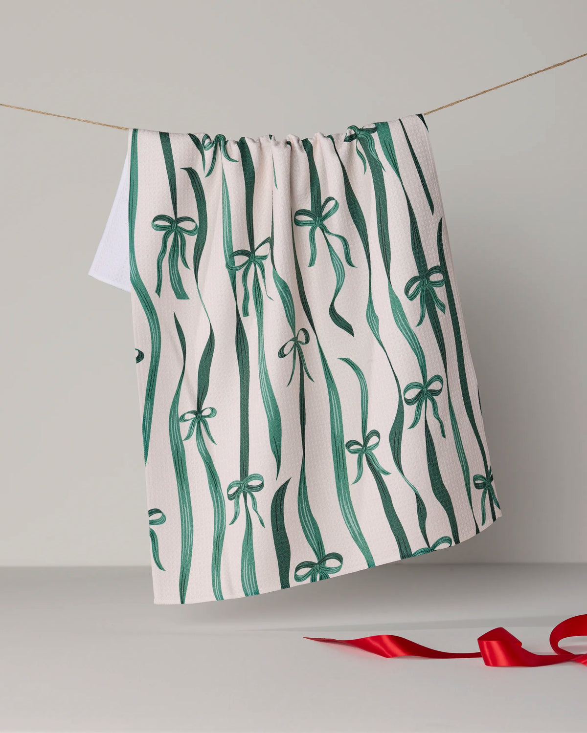 A white, ultra-absorbent Geometry “Falling Ribbon” tea towel featuring a green ribbon and bow pattern hangs on a clothesline, while a red ribbon rests curled on the light gray surface below. The scene is set against a neutral, minimalistic background.