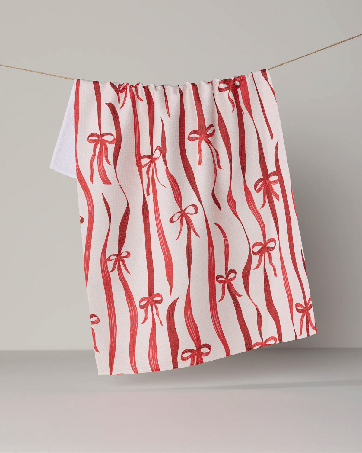 The Geometry “Falling Ribbon” tea towel, featuring a red ribbon and bow pattern on a white background, hangs on a string against a neutral backdrop and is crafted from recycled materials for an eco-friendly touch.