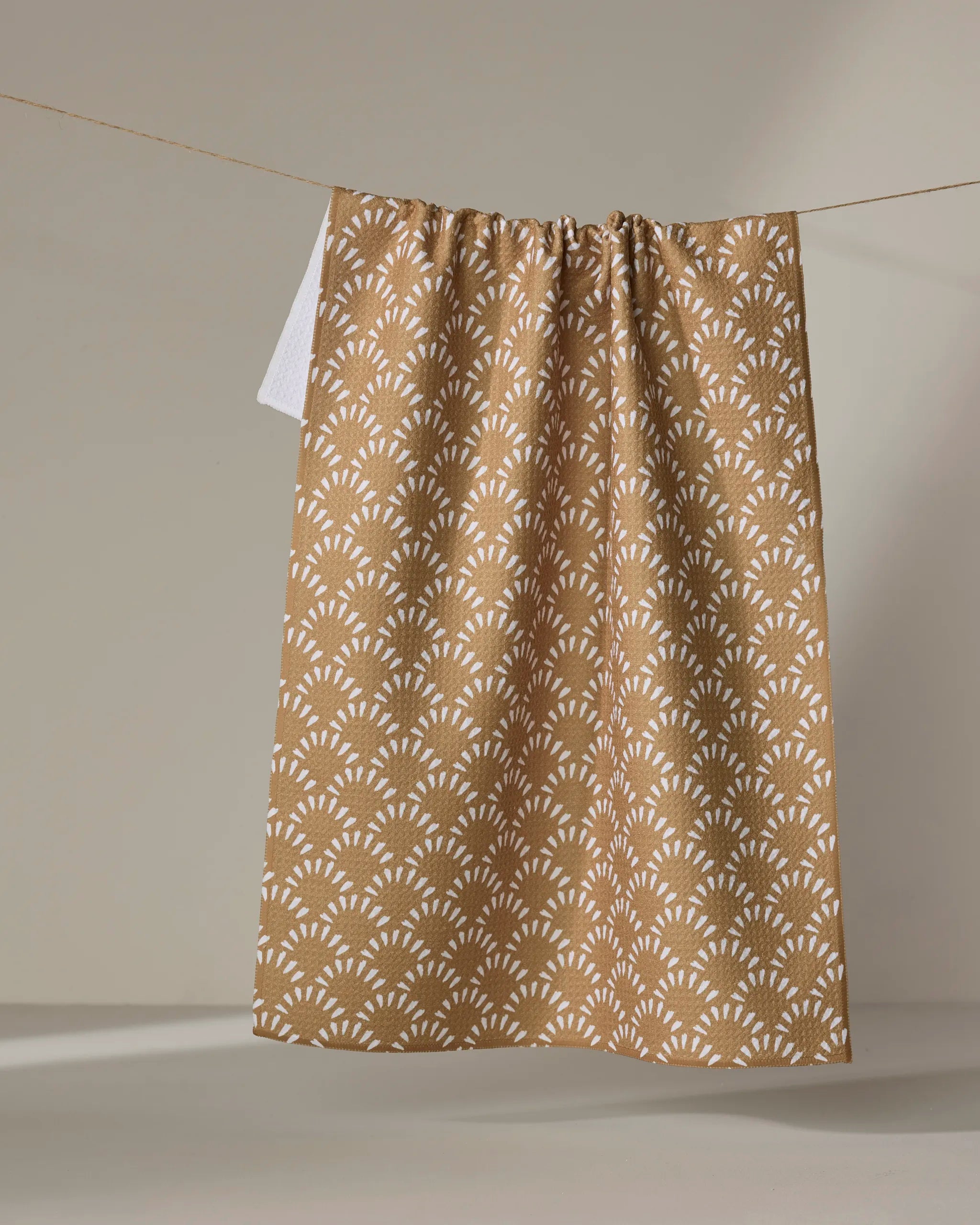 The Fantail kitchen towel by Hufton Studio, made from recycled materials, features a repeating white geometric pattern on brown and hangs on a clothesline against a neutral background.