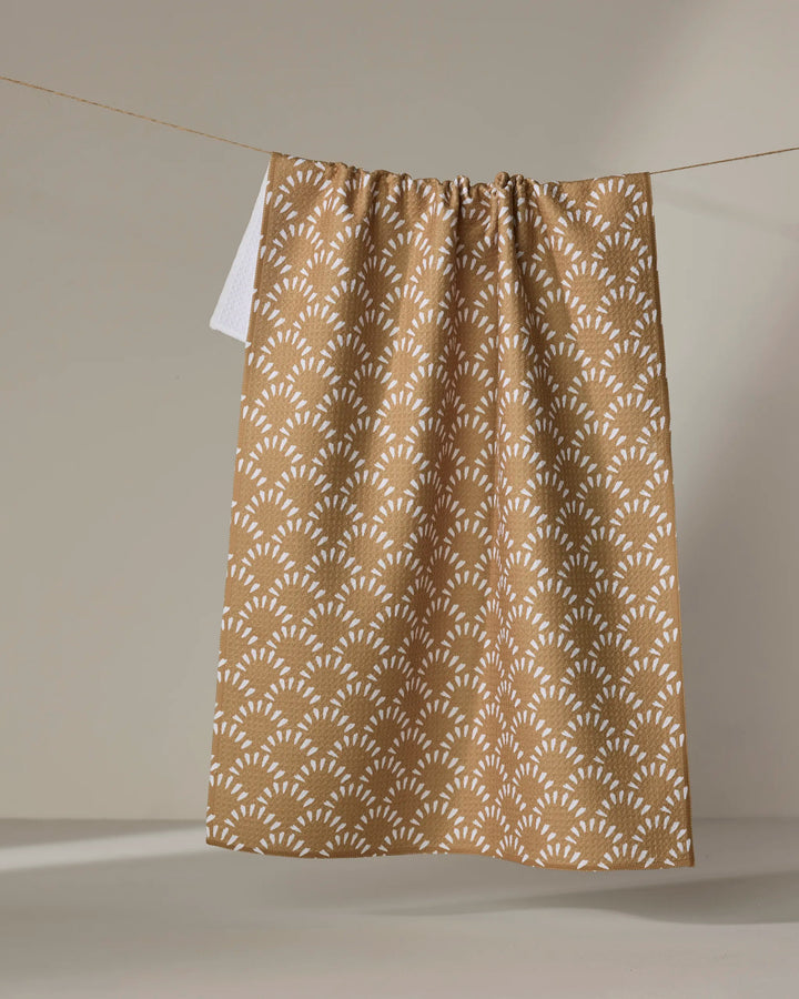 The Fantail kitchen towel by Hufton Studio, made from recycled materials, features a repeating white geometric pattern on brown and hangs on a clothesline against a neutral background.