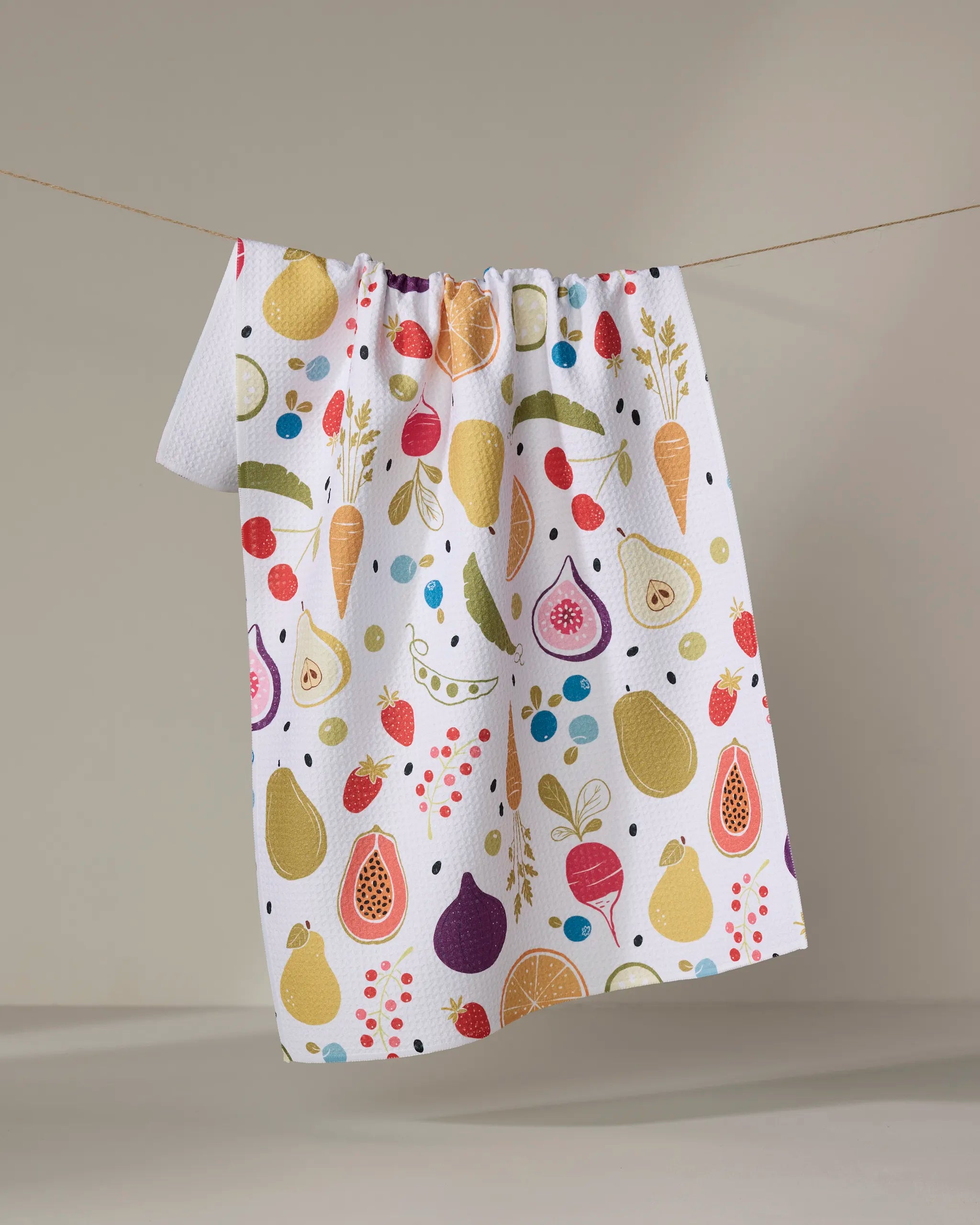 The Sara Mari "Farmers Market" tea towel, ultra-absorbent and white, features a colorful print of pears, figs, berries, peas, and citrus slices. It hangs from a clothesline against a neutral background.