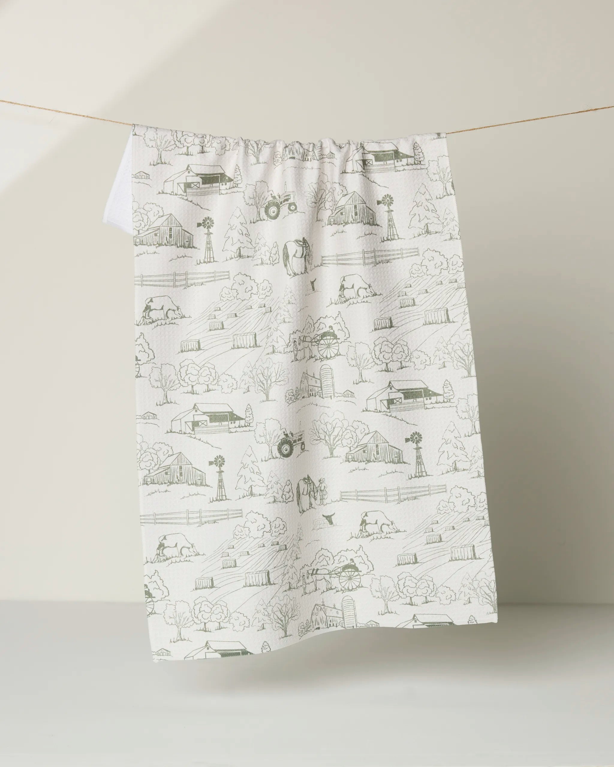 The "Farmyard" tea towel by Hannah Langford is a quick-drying white cloth decorated with green line drawings of rural scenes—houses, trees, animals, and more—hanging on a string against a plain light background.