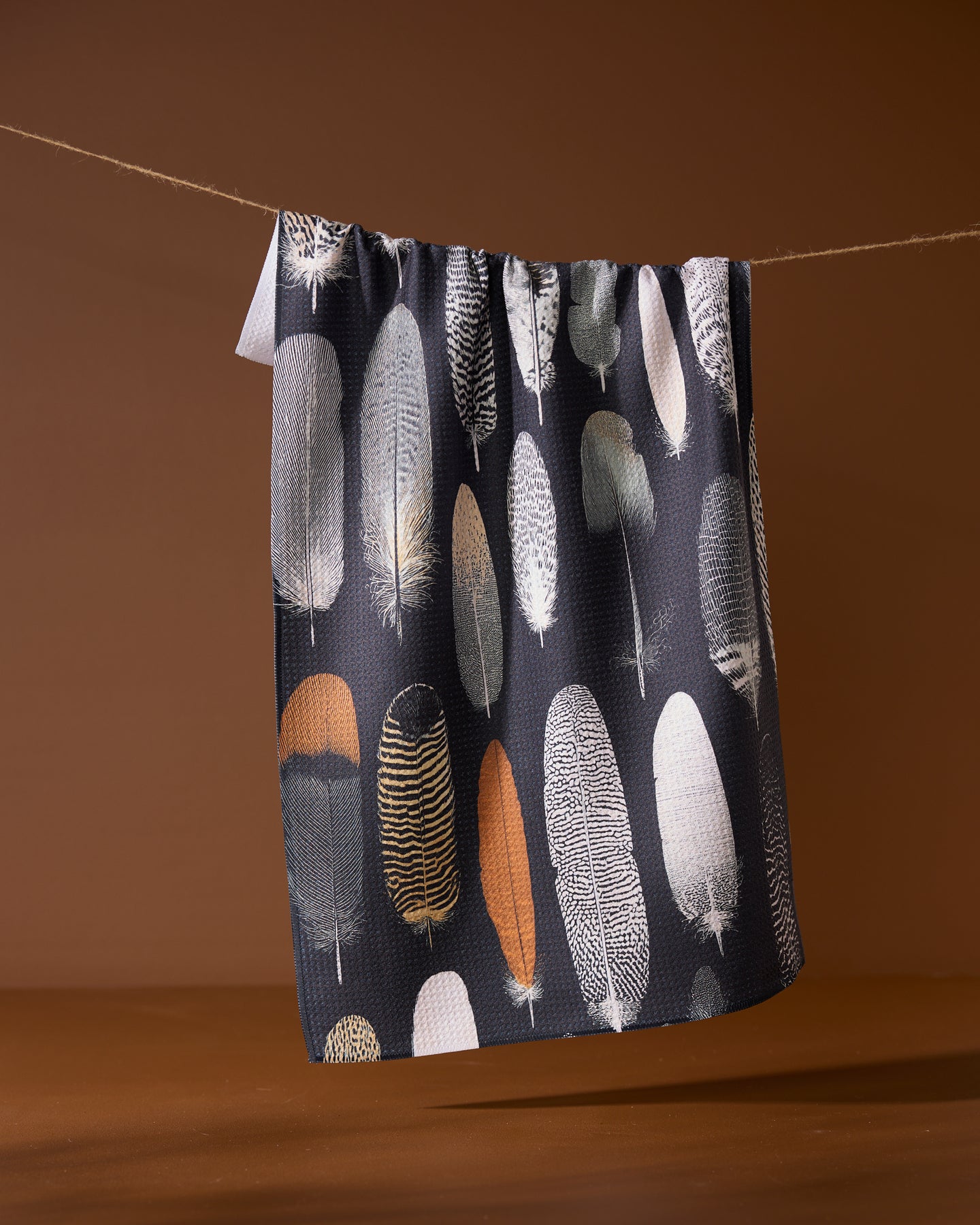 The Geometry Featherfall towel, an eco-friendly, dark-toned piece featuring neutral feather patterns, hangs on a line set against a brown background.
