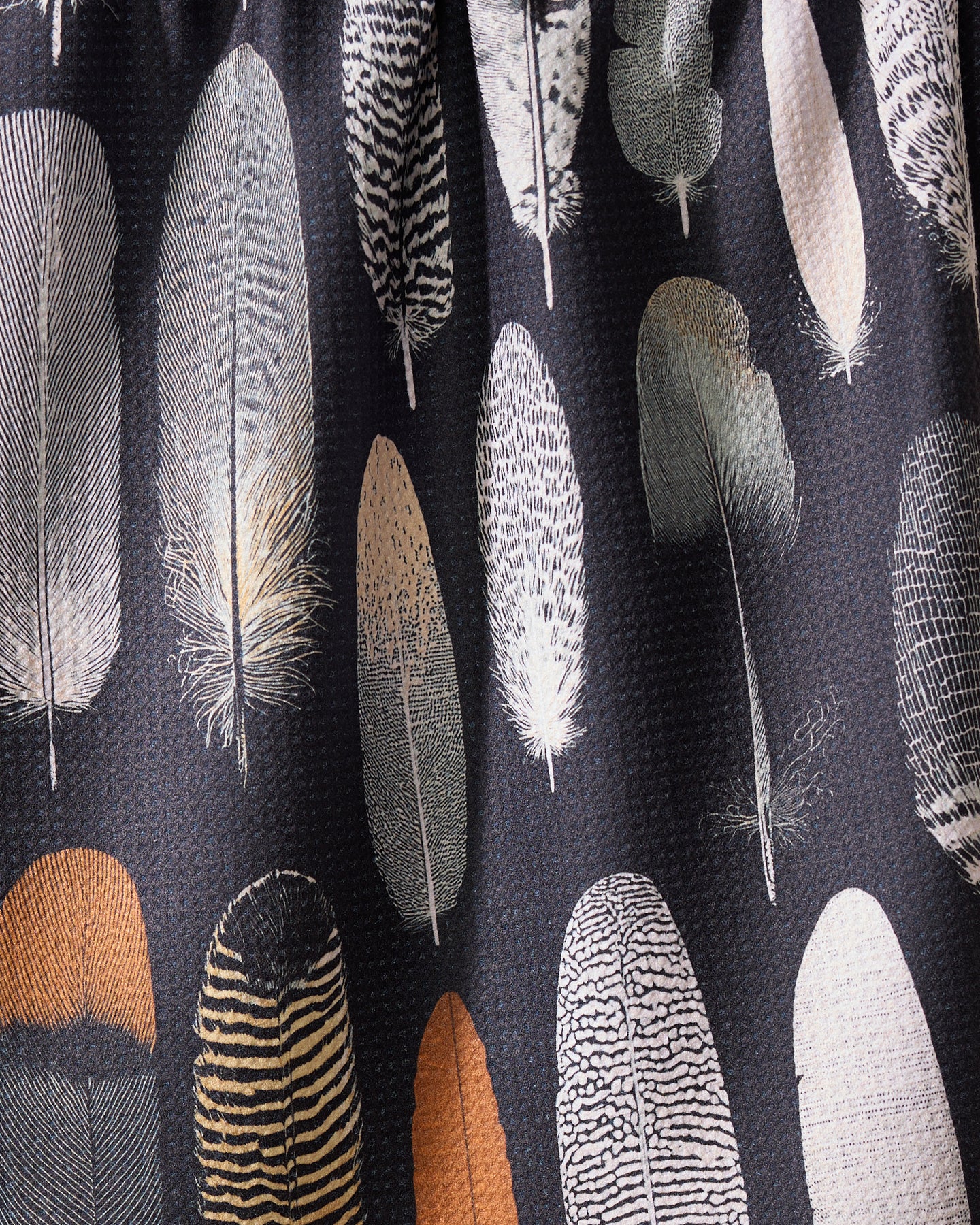 Featherfall by Geometry is an eco-friendly towel with a detailed feather pattern in brown, gray, black, white, and beige shades, featuring colorful feathers arranged vertically on a dark background.