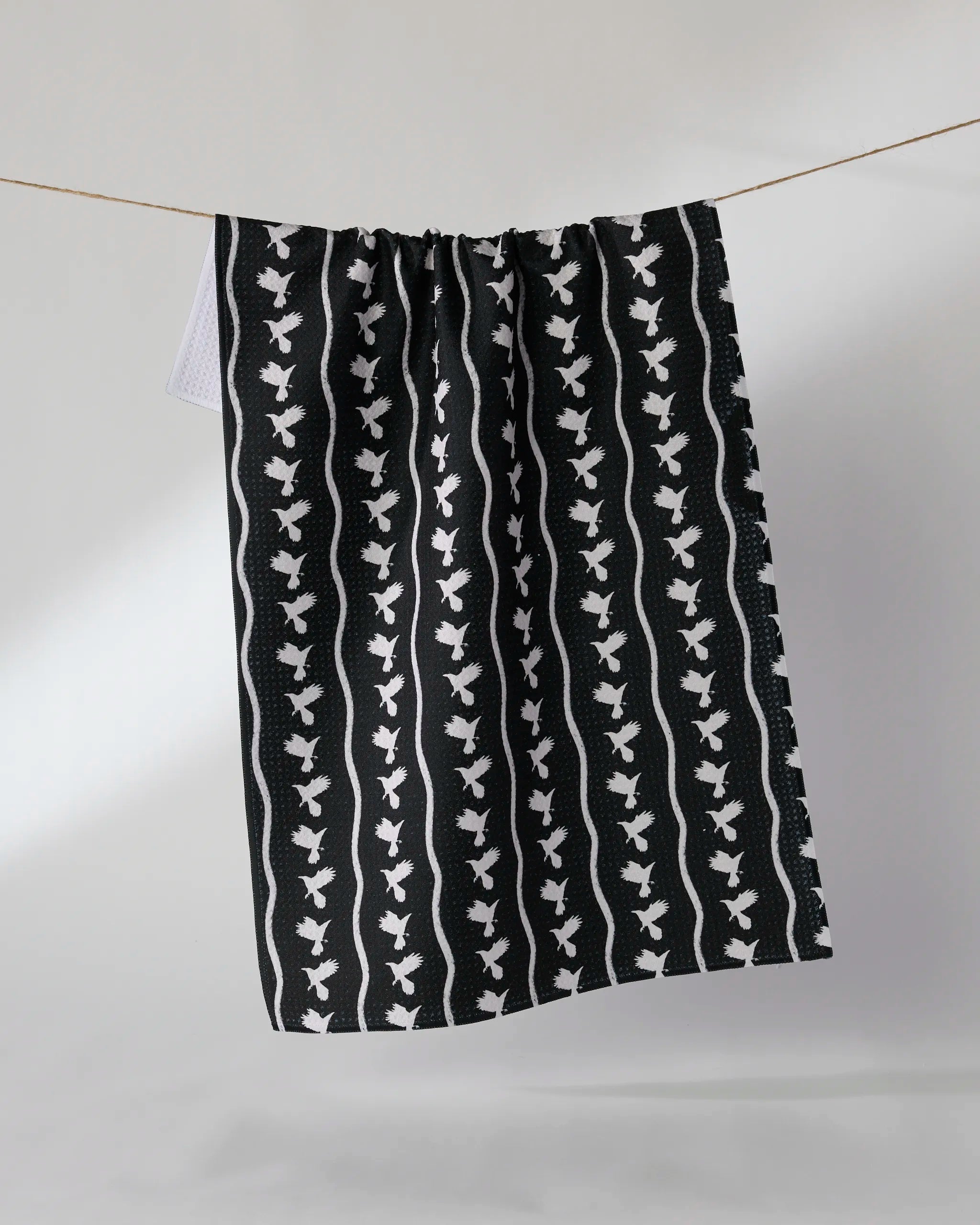 The Geometry Featherline kitchen towel, made from recycled materials, features a black and white bird and wavy line pattern and hangs on a clothesline against a light background.