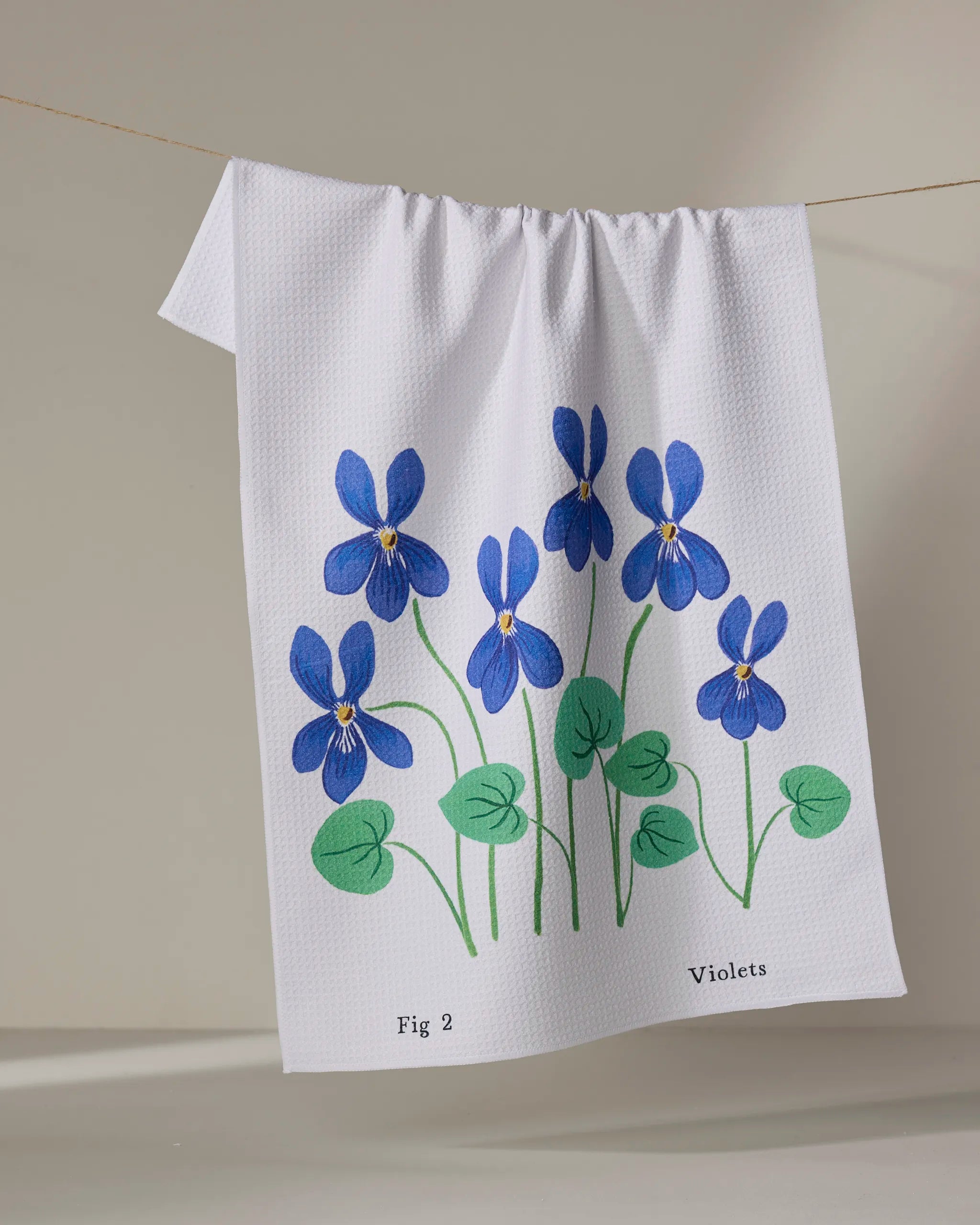 A white tea towel called "February Violets" by Linda Fox features six blue violet flowers and green leaves labeled “Fig 2 Violets,” and is shown hanging on a clothesline against a simple background.