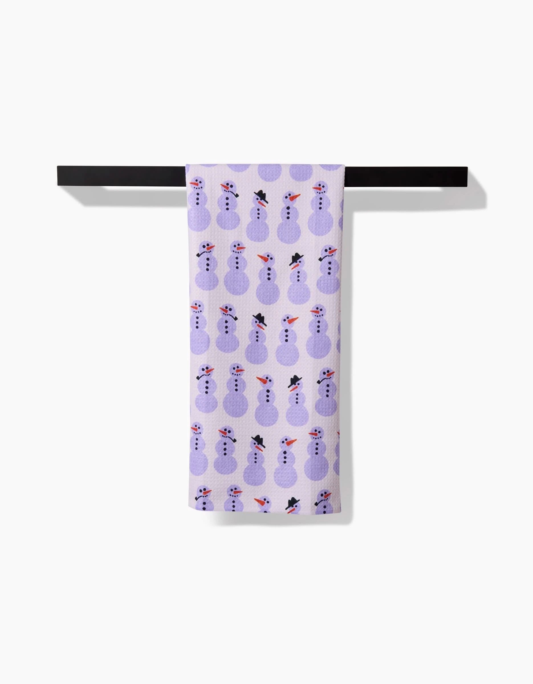 The Amy MacCready Felicity Purple tea towel, featuring a repeating pattern of purple snowmen in black hats and red scarves, hangs neatly from a horizontal black bar against a white background.
