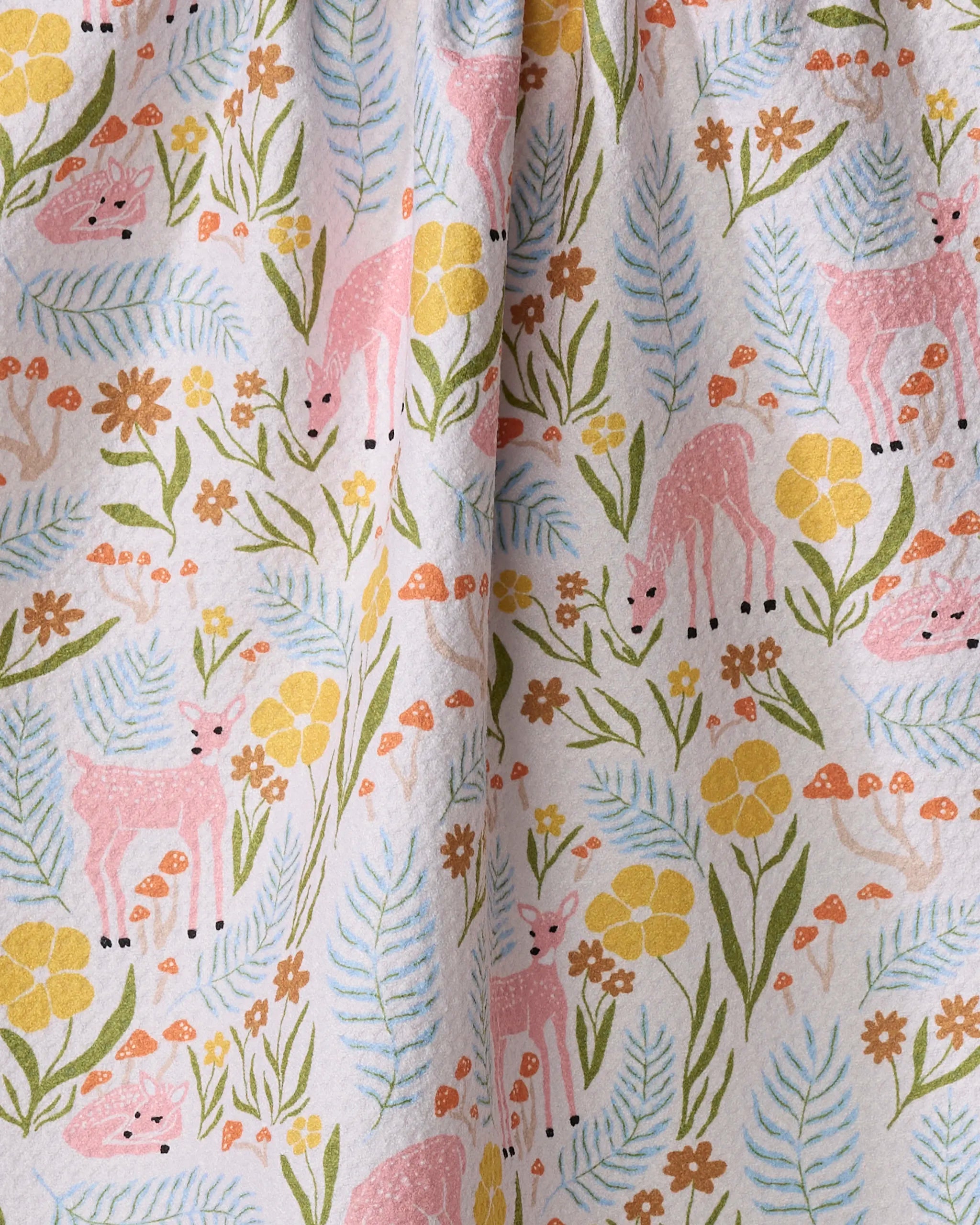 Kayla Ann’s Fern and Fawn kitchen towel is ultra-absorbent and features a whimsical pattern of pink deer, yellow flowers, orange mushrooms, and green leaves on a light background with a vertical center fold.