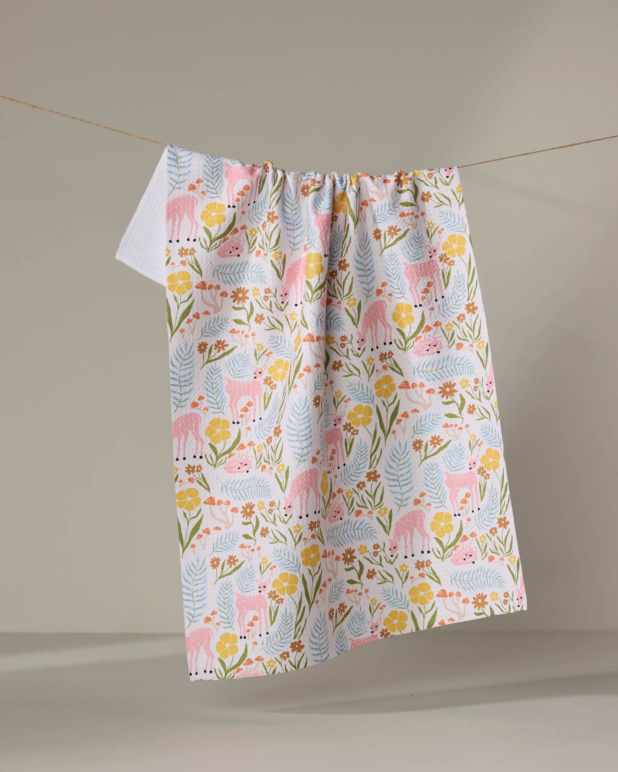 The Kayla Ann "Fern and Fawn" kitchen towel, adorned with pink deer, vibrant flowers, and green leaves, hangs on a clothesline against a beige background.