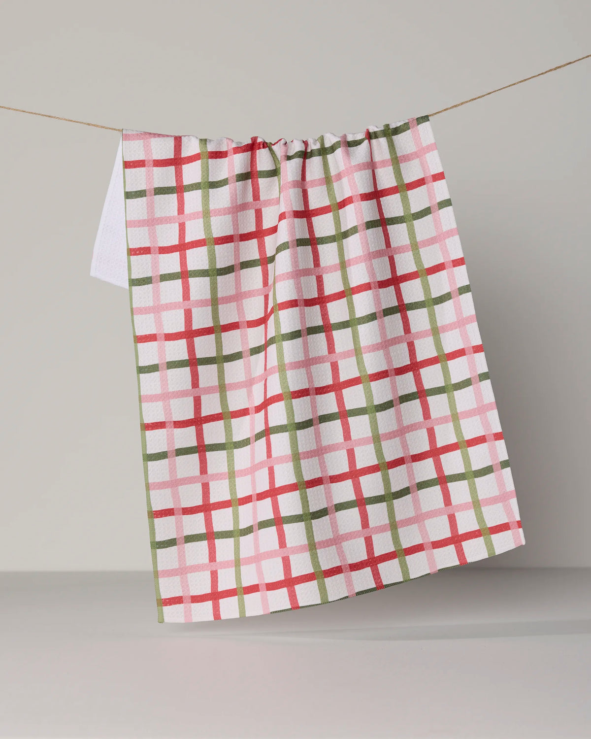 The Geometry Festive Plaid ultra-absorbent tea towel, featuring a red, green, and pink plaid pattern, hangs on a string against a plain light gray background.