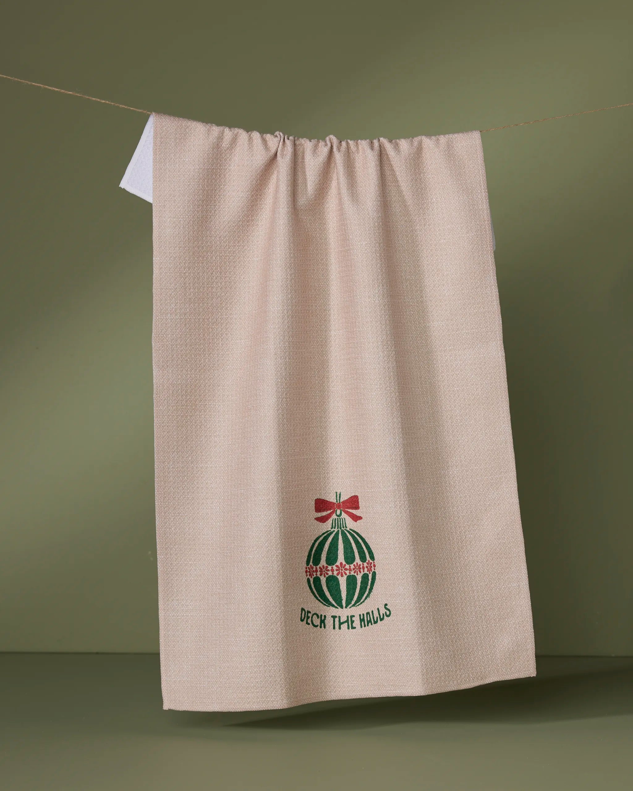 The Geometry Festona tea towel, ultra-absorbent in beige, hangs against an olive green backdrop, embroidered with a Christmas ornament, red bow, and “Deck the Halls” script.