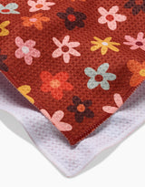 A close-up of the Sara Mari Field Of Fall Daisies kitchen towel, featuring colorful flower patterns on a red background and a white underside—crafted from recycled materials.