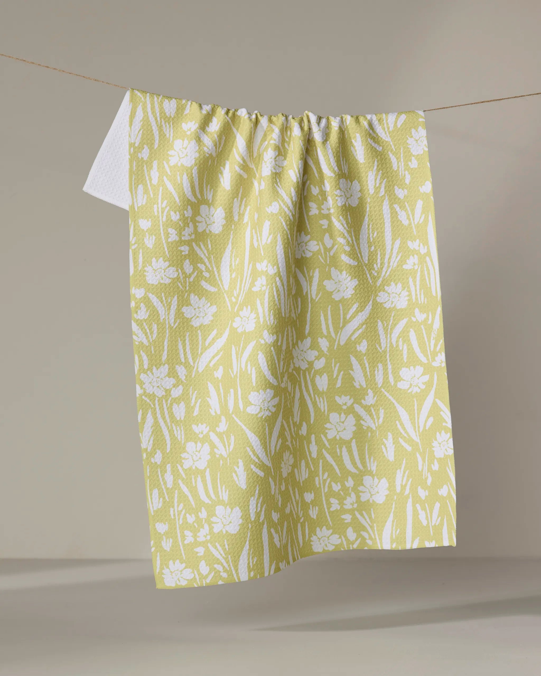 The "Field of Wildflowers" tea towel by Ashley Celena, featuring a white floral pattern on yellow and made from recycled materials, hangs on a clothesline against a neutral background.