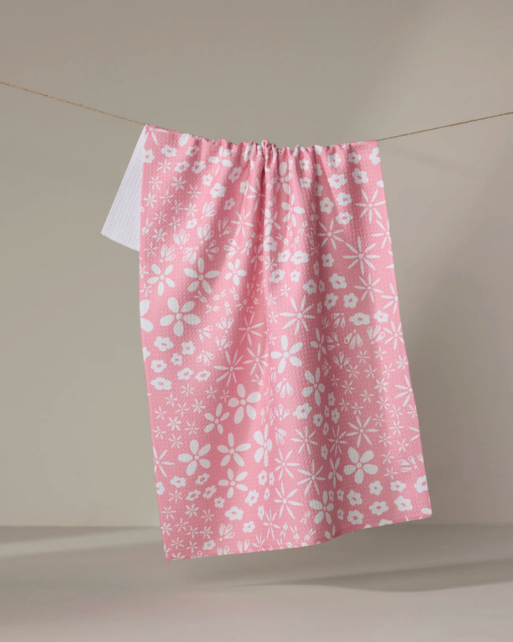 The Fields of Flowers tea towel by Hufton Studio, featuring a pink hue and white floral pattern made from recycled materials, hangs on a clothesline against a beige background with another white cloth partially visible behind it.