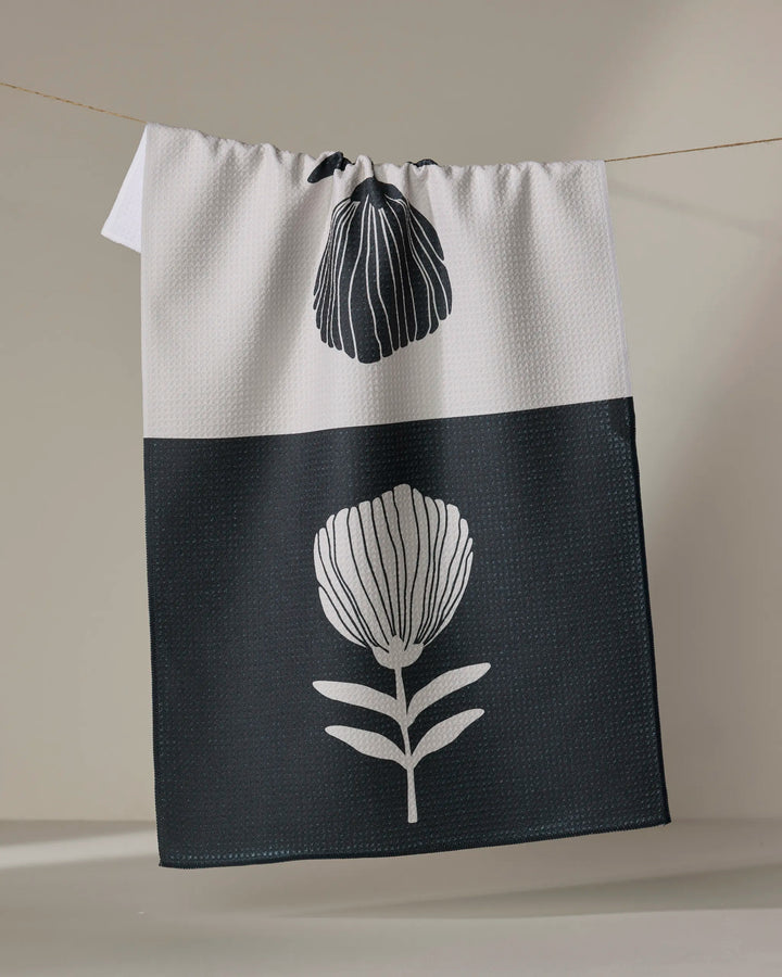 The Fika tea towel by Anna Christian features a minimalist floral design: the top half is white with a black flower bud, and the bottom is black with a large white flower and leaves. Quick-drying, it hangs attractively against any neutral background.